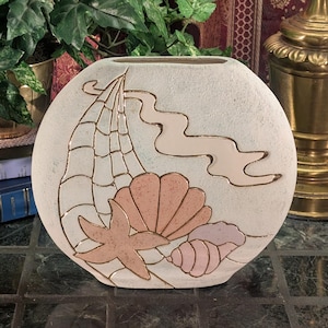 Puede incluir: Un jarrón de cerámica redondo de color blanco roto con un diseño en relieve delineado en oro que muestra una red de pesca, una cinta ondulada, una concha grande en forma de abanico, una estrella de mar y una concha espiral con tonos rosa y melocotón.