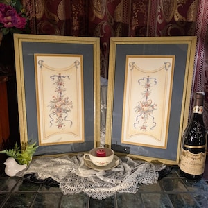 May include: Two framed floral prints with gold frames and blue matting. A teacup with a lit red candle sits on a lace doily. A bottle of wine is on the right, and a white ceramic bird is on the left.