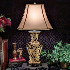 May include: A decorative table lamp with a beige lampshade and a black and gold patterned base. The base features an ornate floral design. The lamp is on a gray surface, with a dark background and other decorative items.