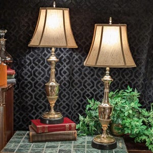 May include: Two brass table lamps with beige fabric shades. The lamps have a classic design with a fluted base and a black circular base. The lamps are placed on a table with books and greenery in the background.