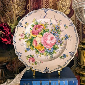 Puede incluir: Un plato decorativo blanquecino con borde festoneado y diseño floral. Las flores pintadas a mano son rosas, amarillas y azules, con hojas verdes. El plato tiene un borde azul y se exhibe sobre un soporte dorado.