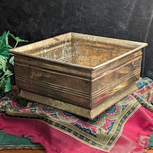 May include: A rectangular brass planter with a slightly aged appearance. The planter has a square shape with a decorative base and horizontal lines. The planter is sitting on a patterned fabric with red and green colors.