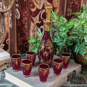 May include: A vintage ruby red decanter set with six matching glasses. The set features gold ornate detailing and a gold stopper. The glasses and decanter are arranged on a book, with a backdrop of a patterned curtain and greenery.