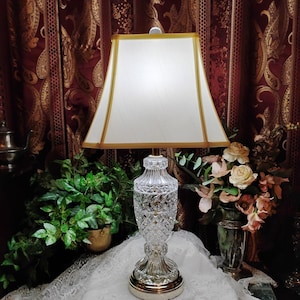 May include: A table lamp featuring a clear, cut-glass base with a diamond-patterned texture and a polished metallic circular bottom. The lamp is topped with a rectangular, off-white fabric shade that has gold-toned trim along the edges.