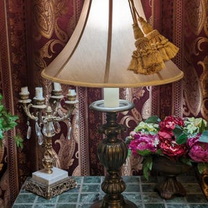 May include: A bronze-colored table lamp with a beige lampshade and decorative tassels. The lamp has a detailed base and is next to a gold candelabra with crystal accents and a floral arrangement. The background features a patterned burgundy curtain.