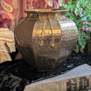Puede incluir: Una maceta de latón con forma hexagonal y textura martillada. El recipiente presenta un borde decorativo de cuerda trenzada alrededor del borde superior y dos detalles de cuerda trenzada que cuelgan por el frente, con acabado metálico.