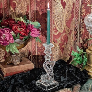 May include: A crystal candlestick with a mermaid design holds a green candle. The candlestick is on a black velvet surface with a book and a vase of flowers in the background.