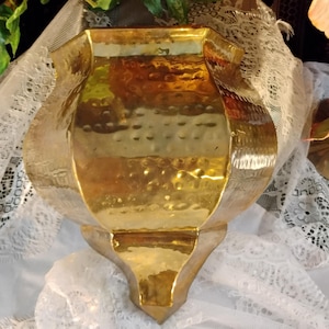 Puede incluir: Una maceta de pared de latón pulido en color dorado con una abertura superior ancha y acampanada y una base puntiaguda y estrecha. La superficie del recipiente de metal muestra una textura martillada distintiva, creando un aspecto reflectante.