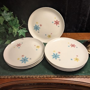 May include: A set of white ceramic dinner plates featuring a scattered pattern of starburst-shaped motifs in shades of blue, red, and yellow. The plates are arranged in two stacks with one plate standing upright behind them.
