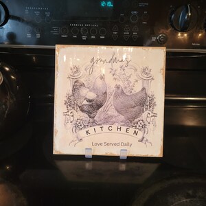 May include: A white ceramic sign with a black and white illustration of two chickens and floral accents. The sign reads "grandma's kitchen Love Served Daily".