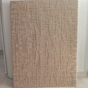 May include: A rectangular panel with a woven texture in shades of beige and tan. The pattern is a tight weave, creating a grid-like design. The panel is standing upright against a white wall.