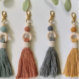 May include: Four handmade macrame keychains with gold-colored clasps. Each keychain features a tassel in different colors: teal, rust, gray, and mustard yellow. They also have wooden rings and decorative beads.