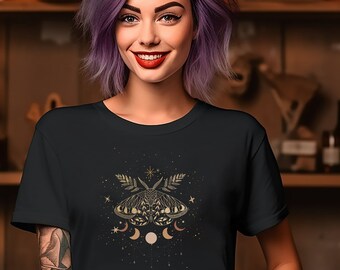 May include: A black t-shirt featuring a gold moth and celestial design. The graphic includes a moth, crescent moons, stars, and foliage. The person in the image is wearing the t-shirt and smiling.