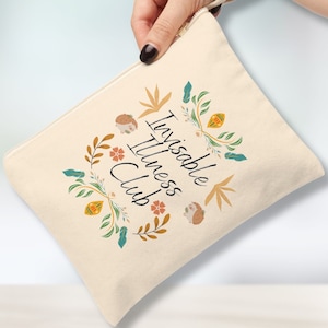 May include: Cream-colored canvas pouch with a wrist strap, featuring the text "Invisible Illness Club" surrounded by floral and hedgehog illustrations in orange, teal, and brown. The pouch has a zipper closure.