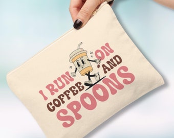 Pode incluir: Bolsa de lona creme com fecho de correr e alça de pulso. Apresenta um gráfico retro de uma chávena de café numa prancha de surf com o texto "I RUN ON COFFEE AND SPOONS" em rosa e castanho.