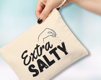 Pode incluir: Uma bolsa de lona bege com um gráfico preto e branco de uma mão espalhando sal. O texto "Extra Salty" está impresso em preto na bolsa.