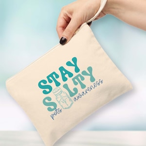 Peut inclure: Une pochette zippée en toile beige avec un design dégradé bleu et vert. La pochette a une sangle blanche et le texte "Stay Salty" avec un dessin de salière et les mots "pots awareness".