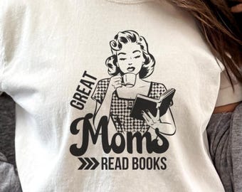 Op de afbeelding: Een wit T-shirt met een zwart-witte illustratie van een vrouw die een boek leest en koffie drinkt. De tekst op het shirt luidt "Great Moms Read Books".