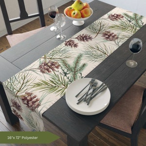 Könnte beinhalten: Ein beiger Tischläufer mit einem Aquarelldesign aus Kiefernästen und Tannenzapfen. Der Läufer ist 16 Zoll breit und 72 Zoll lang und besteht aus Polyester.
