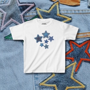 May include: White t-shirt with a design of denim stars in shades of blue. The stars are stitched onto the shirt. The text "Graceful Affirmations" is printed on the upper right of the shirt.