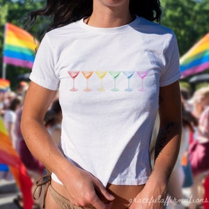 Girl wearing pride baby tee in front of pride parade. The baby tee features red, orange, yellow, green, blue, and purple martinis