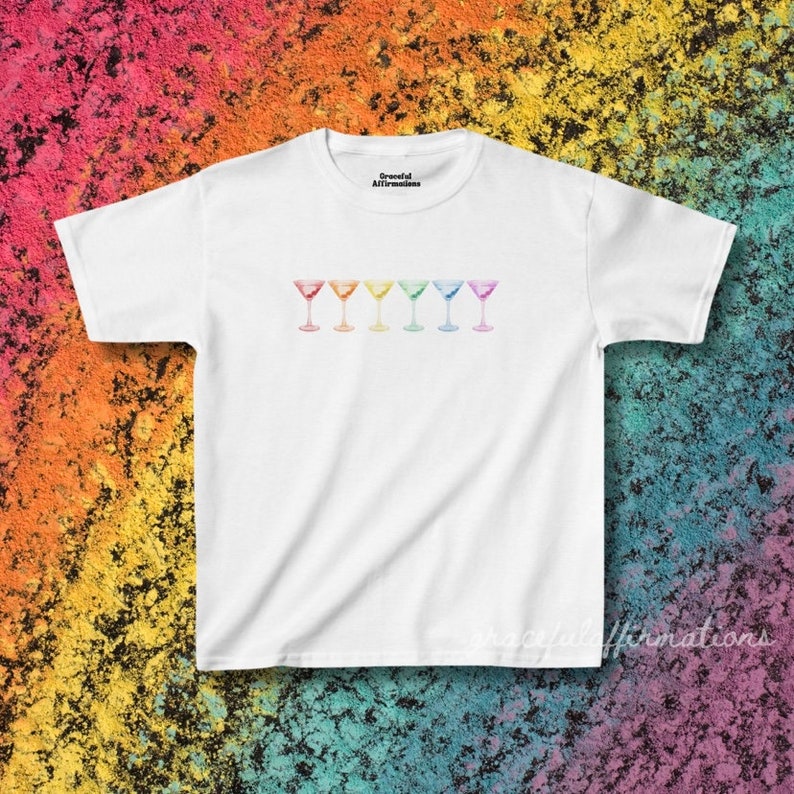 White version of the baby tee in front of rainbow chalk background. The baby tee features rainbow martini glasses