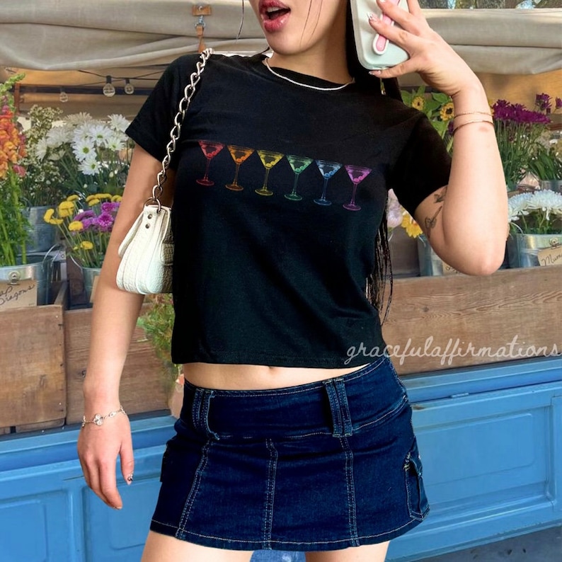 Girl wearing black pride baby tee with rainbow martinis, with a short denim skirt