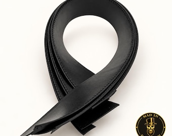 May include: A black leather belt strap featuring a smooth finish and detailed edge stitching. The strap is coiled into a loop shape. In the bottom right corner, there is a circular logo with a skull wearing a top hat and the text MAD IN BROOKLYN.