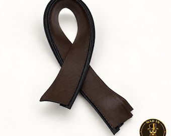 May include: A dark brown leather strip folded into a ribbon shape with black stitched edges. The leather has a smooth matte finish and is curved into a loop. In the bottom right corner, a circular logo features a skull wearing a top hat with the text MAD IN BROOKLYN.