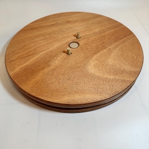 May include: A round wooden turntable with two dowel rods in the center. The turntable is made of light brown wood and has a smooth surface.