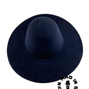 May include: A navy blue fedora hat with a wide brim. The hat has a rounded crown and a circular brim. The hat is made of a soft material. The text "MAD IN BROOKLYN" is printed on the lower right side of the image.