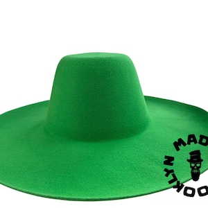 May include: A bright green hat with a wide brim and a cylindrical crown. The hat has a felt-like texture and features the text "MAD IN BROOKLYN" in a circular design with a skull logo. The hat is set against a white background.