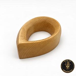 May include: A smooth, light brown wooden object shaped like a teardrop with a hollow center. The wood grain is visible across the curved surface. In the bottom right corner, a circular logo features a skull wearing a top hat with the text MAD IN BROOKLYN.
