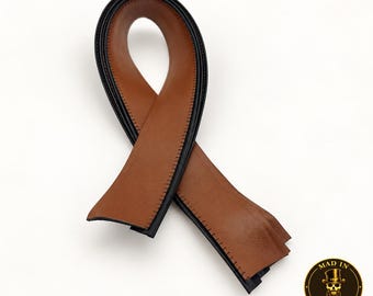 May include: A tan leather strap folded into a loop, featuring black edges and visible stitching along the sides. The strap has a smooth texture and a consistent width throughout its length. A circular logo with a skull in a top hat reads MAD IN BROOKLYN.