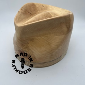 May include: A wooden hat block with a smooth, light brown finish. The block is shaped like a traditional fedora hat. The bottom of the block is made of plywood and has the text "MADE IN BROOKLYN" printed on it.
