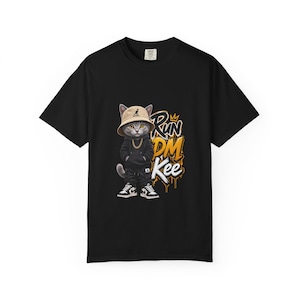 Może przedstawiać: Czarny t-shirt z krótkim rękawem z grafiką kota w beżowym kapeluszu typu bucket hat, czarnej bluzie z kapturem i złotym łańcuchu. Kot stoi obok stylizowanego żółto-białego napisu RUN DM Kee z małą ikoną korony umieszczoną nad tekstem.