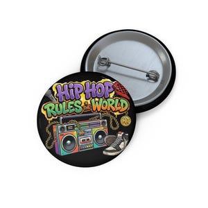May include: A round pin-back button featuring a colorful illustration of a boombox, a high-top sneaker, and a microphone. The text reads Hip Hop Rules The World in stylized graffiti lettering. The background is black with gold chain accents.
