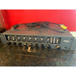 May include: A gray Peavey rack-mount audio mixer featuring a series of black and white control knobs on the front panel, a power button, and a black power cord with a three-prong plug resting on top of the unit. The text Peavey Made in U.S.A. is visible.