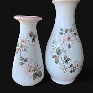 May include: Two white ceramic vases with flared rims and a pink trim. Each vase features a botanical illustration of light pink flowers with yellow centers, green leaves, and brown stems. The vases have a smooth, glossy finish and curved bodies.