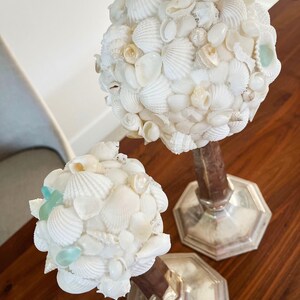 May include: Two decorative seashell spheres atop silver candle holders. The spheres are composed of various white seashells, with a few pieces of sea glass. The candle holders have a brown wooden stem and a silver base.