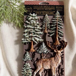 Puede incluir: Un molde decorativo de silicona marrón titulado Stag of the Highlands que presenta diseños en relieve de un ciervo de pie con grandes astas, varios pinos de diferentes tamaños y patrones de follaje de helecho. El empaque incluye rayas rojas y blancas.