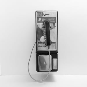 Op de afbeelding: Een zwarte en zilveren telefooncel met een zwarte draaischijftelefoon en een kabel die eraf hangt. De telefoon heeft een bord met de tekst "Save Money Use Coin Calls 50c".
