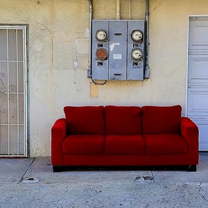 Peut inclure: Un canapé rouge est placé devant une porte de garage blanche et une porte blanche avec un portail métallique. Le canapé a trois coussins et se trouve devant un mur blanc.
