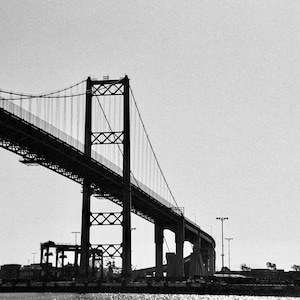 May include: A black and white photo of a large bridge with a steel structure and cables. The bridge is over water and there are buildings in the distance.