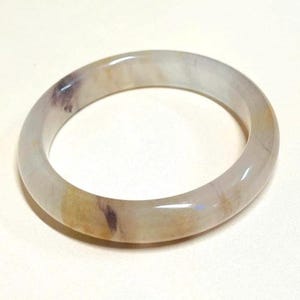 May include: A translucent jade bangle bracelet. The bracelet features a mix of colors, including white, yellow, and purple. The smooth, polished surface reflects light, highlighting the natural variations in the stone. A classic piece of jewelry.