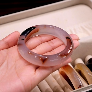 May include: A smooth, translucent bangle bracelet crafted from polished agate stone. The circular band features a pale, milky white base color with distinct, dark brown and black natural inclusions scattered throughout the polished surface.