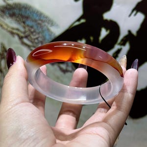 May include: A translucent agate bangle bracelet. The bracelet has a colour gradient, shifting from clear, milky white to a vibrant orange-red hue. The polished surface reflects light, highlighting the natural patterns within the stone.