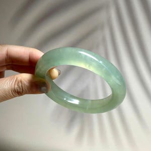 Puede incluir: Un brazalete tipo esclava de jade verde claro, translúcido y liso, con una superficie pulida y redondeada. El accesorio circular presenta sutiles variaciones en la intensidad del color y la claridad, mostrando un diseño de banda sólida y continua.
