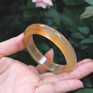 Może przedstawiać: Gładka, półprzezroczysta bransoletka typu bangle w ciepłej, miodowo-żółtej i bursztynowej kolorystyce. Okrągły dodatek posiada polerowane, błyszczące wykończenie, które odbija światło, ukazując subtelne wewnętrzne różnice w odcieniu materiału.