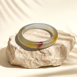 May include: A translucent jade bangle bracelet with a gradient of colors, including yellow, gray, and a touch of red. The bracelet is resting on a textured, light-colored stone. This piece is a beautiful example of natural gemstone jewelry.
